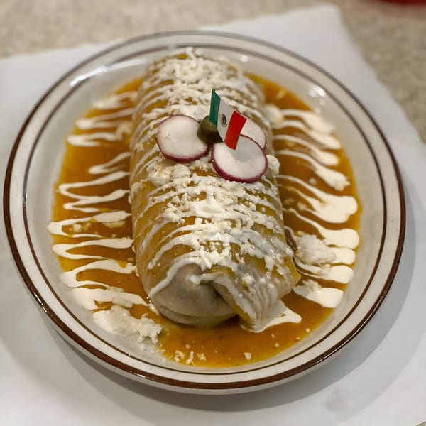 A large burrito covered in orange sauce, white cream, and cheese, garnished with radishes and a Mexican flag.