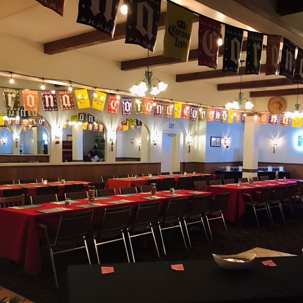 A large dining room at Las Cazuelas Restaurant with red-clothed tables, chairs, and festive Corona banners.
